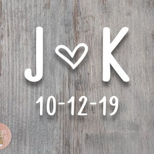 May include: A white decal featuring the letters J and K separated by a heart symbol, with the date 10-12-19 printed below in a clean sans-serif font. The design is displayed against a weathered gray wood grain background texture.