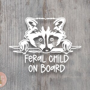 May include: A white vinyl decal featuring a stylized raccoon peering over a horizontal line with its paws visible. Below the raccoon, the text reads FERAL CHILD ON BOARD in a casual, hand-lettered font. The design is presented as a single graphic element.