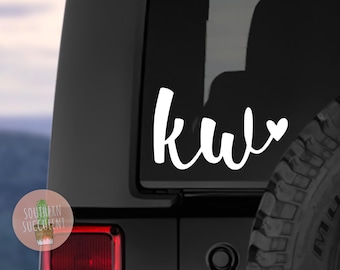 May include: A white vinyl decal applied to a dark vehicle window, featuring the lowercase letters kw in a flowing script font followed by a small heart symbol. The design is positioned on the rear side glass of a black sport utility vehicle.