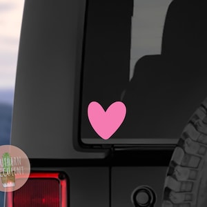 May include: A solid pink heart-shaped decal applied to the rear window of a black vehicle. The heart is a simple, symmetrical graphic design with smooth, rounded edges, positioned centrally on the dark glass surface of the car window.