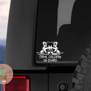 May include: White vinyl decal of a raccoon peeking over a horizontal banner with the text 'Feral Children On Board'. The decal is affixed to the rear of a black Jeep. The Jeep's tire is partially visible in the lower right corner of the image. The decal is a humorous design suitable for parents of mischievous children.