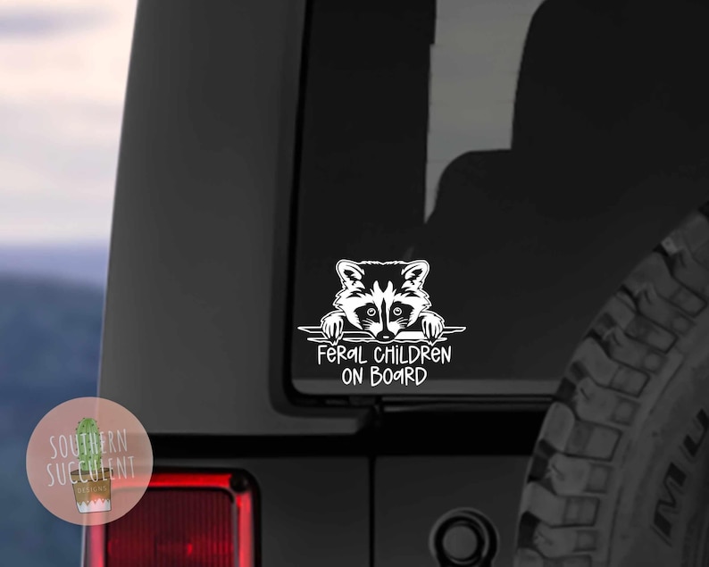 May include: White vinyl decal of a raccoon peeking over a horizontal banner with the text 'Feral Children On Board'. The decal is affixed to the rear of a black Jeep. The Jeep's tire is partially visible in the lower right corner of the image. The decal is a humorous design suitable for parents of mischievous children.