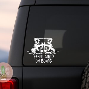 May include: A white vinyl decal applied to a dark vehicle window, featuring a graphic illustration of a raccoon peering over a horizontal line, with the text FERAL CHILD ON BOARD printed in a bold, sans-serif font directly underneath the raccoon.