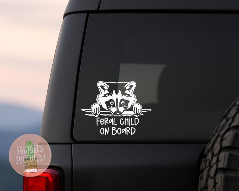 May include: A white vinyl decal applied to a dark vehicle window, featuring a graphic illustration of a raccoon peering over a horizontal line, with the text FERAL CHILD ON BOARD printed in a bold, sans-serif font directly underneath the raccoon.
