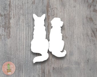 May include: White silhouette of two dogs sitting side by side. The dog on the left is a larger breed with a long tail, while the dog on the right is a smaller breed with a shorter tail. The silhouettes are cut out of a white material and placed on a wood background.