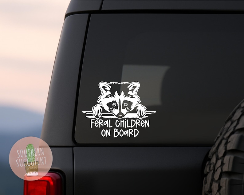 May include: A white vinyl decal applied to a dark vehicle window featuring a graphic illustration of a raccoon peering over a horizontal line with the text FERAL CHILDREN ON BOARD printed in a bold, sans-serif font underneath the animal.