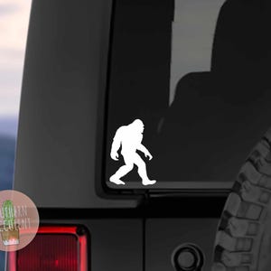 May include: A white vinyl decal featuring the silhouette of a walking Bigfoot creature, applied to the rear window of a dark-colored vehicle. The design shows the iconic bipedal figure in profile, facing right, with a distinct hairy appearance.