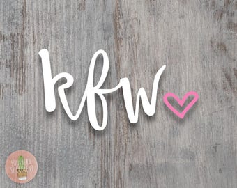 May include: A white script decal featuring the letters kfw in a lowercase cursive font, accompanied by a small pink hand-drawn heart symbol positioned to the right of the text. The design is presented against a weathered gray wood grain background.