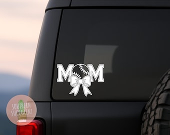 May include: A white vinyl decal applied to a vehicle window featuring the text MOM in a block font. The letter O is replaced by a white graphic of a baseball with visible stitching, positioned directly above a large white decorative bow.