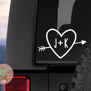 May include: White vinyl decal of a heart with an arrow through it. The heart has the initials "J+K" inside. The decal is on a black car.