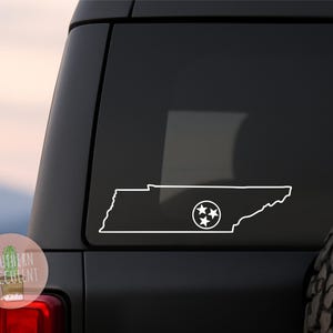 May include: A white vinyl decal featuring the outline of the state of Tennessee with the three-star emblem from the state flag positioned on the right side, applied to the rear window of a dark-colored vehicle with a spare tire visible.