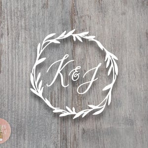 May include: A white vinyl decal featuring the letters K & J in a cursive script font, enclosed within a circular wreath of stylized leaves. The design is presented against a gray wood grain background, with a small circular logo in the corner.