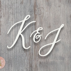 May include: A white cutout sign featuring the letters K and J connected by an ampersand in a flowing cursive script font. The letters are positioned against a weathered gray wood grain background with a small circular logo in the bottom corner.