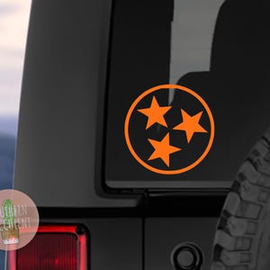 May include: A bright orange vinyl decal featuring three five-pointed stars arranged within a circular border. The design is applied to the dark tinted rear window of a vehicle, positioned above the red taillight and next to a spare tire.