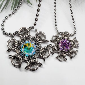 May include: Two silver necklaces with snowflake-shaped pendants. The pendant on the left has a teal iridescent crystal in the centre. The pendant on the right has a purple iridescent crystal in the centre.
