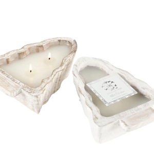 May include: Two-wick candle in a distressed, white-washed wooden container. The candle has a rectangular shape with a natural, organic edge. A label is visible on the second candle, with text and a floral design. The candle is a home decor item.