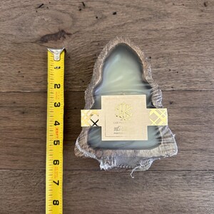 May include: A candle in a wooden holder, approximately 4 inches wide, is shown. The candle is wrapped in clear plastic and has a gold and white label. A yellow tape measure is next to the candle.