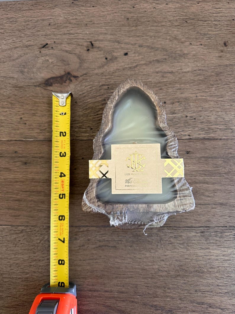 May include: A candle in a wooden holder, approximately 4 inches wide, is shown. The candle is wrapped in clear plastic and has a gold and white label. A yellow tape measure is next to the candle.