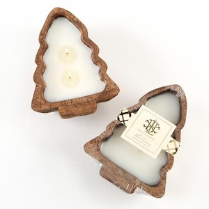 May include: Two Christmas tree-shaped candles with a wooden base. The candles have a white wax and two wicks. One candle has a tag attached with a black and gold ribbon. The candles are on a white surface.