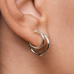 Könnte beinhalten: Silberne Creolen mit einem Doppel-Creolen-Design. Die Ohrringe werden am Ohr einer Person mit dunklen Haaren getragen.