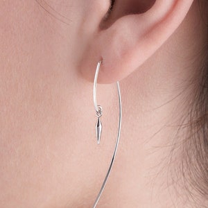 May include: A silver earring featuring a long, curved bar and a small, pointed charm. The earring is worn in the ear, with the bar extending downwards. The charm hangs from a small loop.