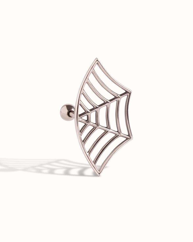 May include: A silver-toned metal ear stud featuring a flat, fan-shaped spider web design. The jewellery piece includes a straight post with a small, round ball backing, showcasing a detailed, open-work web pattern with curved outer edges.