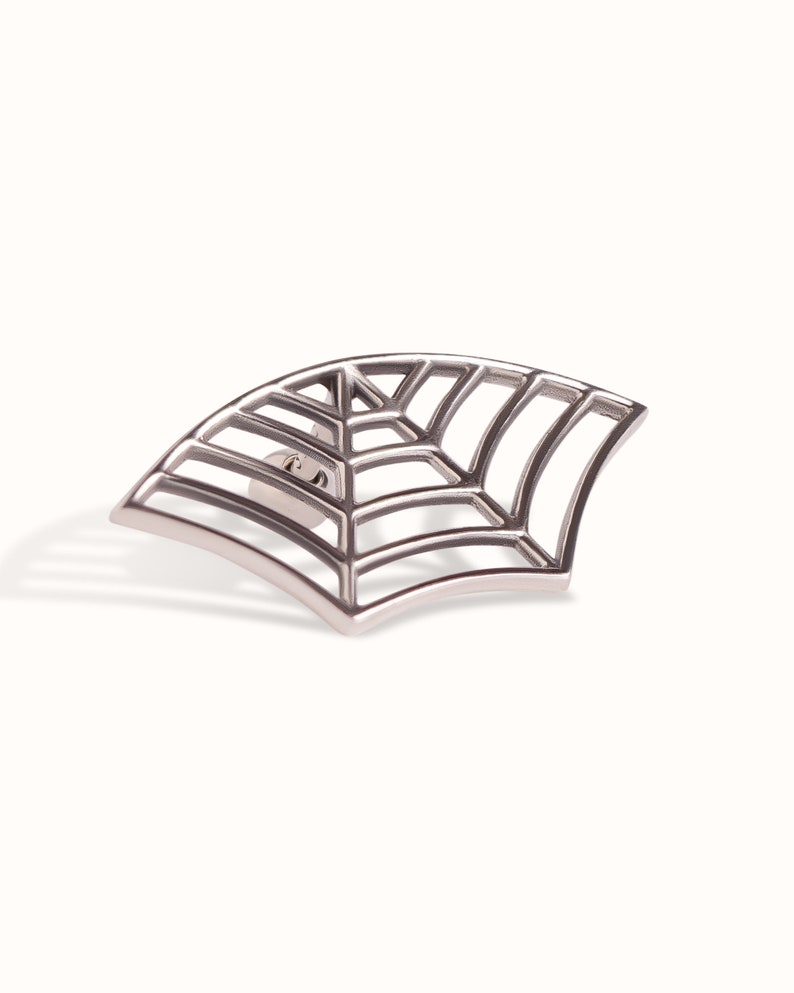 May include: A silver-toned metal pin shaped like a segment of a spider web. The design features a series of curved, radiating lines intersected by concentric arcs, creating a geometric grid pattern with a polished, metallic finish and a pin back.