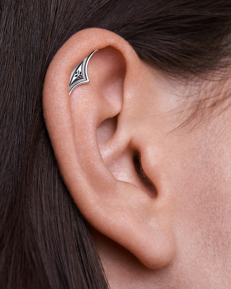 May include: A silver ear cuff with a floral design. The cuff is curved and has a pointed end. The floral design is intricate and detailed.