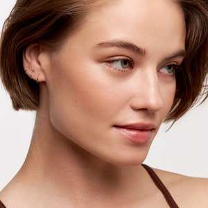 May include: A close-up of a woman's face with short brown hair. She is wearing three small gold hoop earrings in her left ear.