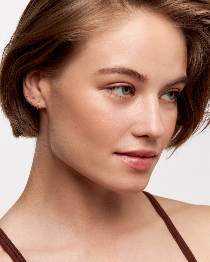 May include: A close-up of a woman's face with short brown hair. She is wearing three small gold hoop earrings in her left ear.