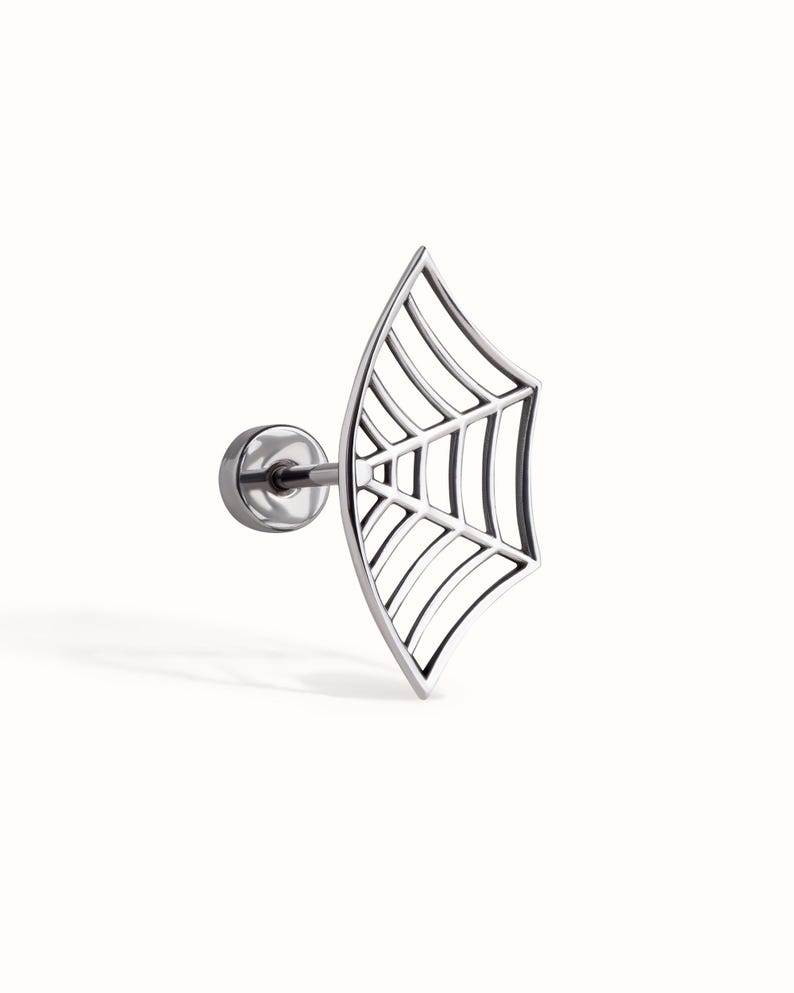 May include: A silver-toned stud earring featuring a flat, open-work spider web design. The piece includes a circular backing post and a curved, fan-shaped web motif with thin metal lines radiating from a central point to the outer edge.