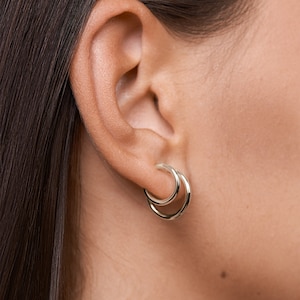 Puede incluir: Un par de pendientes de aro de plata con un dise&ntilde;o de aro doble. Los pendientes se llevan en la oreja de una persona.