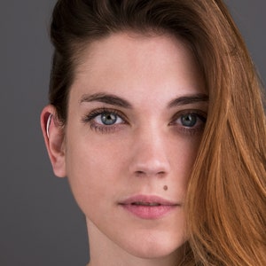 May include: A woman with long, wavy brown hair. She is wearing a black shirt and has a mole on her lower lip. Her eyes are blue and she is looking directly at the camera.