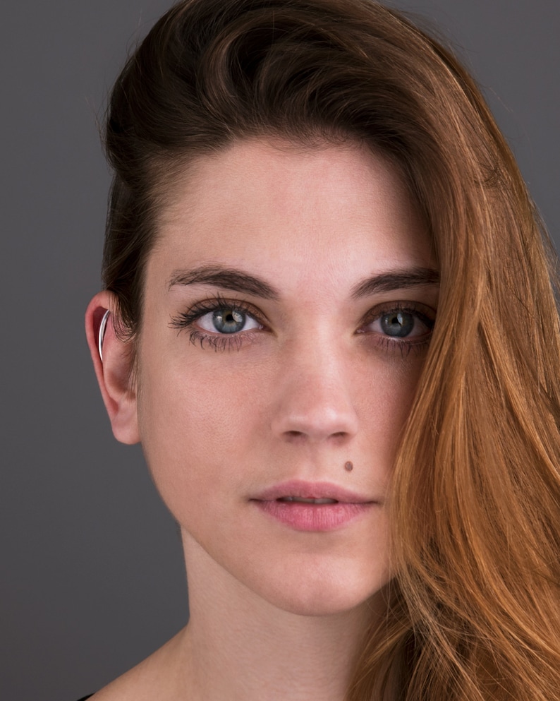 May include: A woman with long, wavy brown hair. She is wearing a black shirt and has a mole on her lower lip. Her eyes are blue and she is looking directly at the camera.