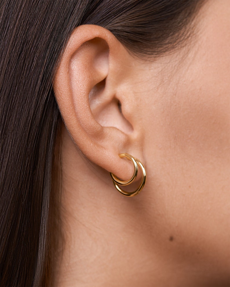 Puede incluir: Un par de pendientes de aro de oro usados en la oreja de una persona. Los pendientes son aros dobles, con un aro ligeramente m&aacute;s grande que el otro.