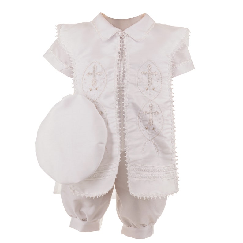 May include: White christening outfit for a baby boy, featuring a short-sleeved jacket with embroidered crosses, matching pants, and a white hat.