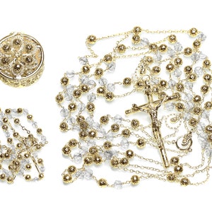 May include: A gold-toned rosary with clear crystal beads and a crucifix. The rosary is laid out on a white background. There is a small gold-toned box with a floral design in the upper left corner of the image.