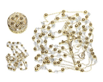 May include: A gold-toned rosary with clear crystal beads and a crucifix. The rosary is laid out on a white background. There is a small gold-toned box with a floral design in the upper left corner of the image.