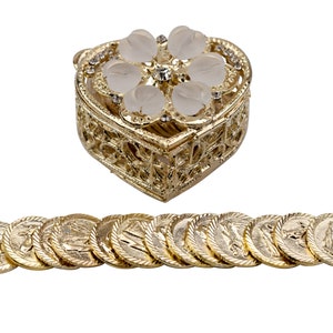 Puede incluir: Una caja de corazón dorada con un diseño de flor blanca y un centro de cristal transparente. La caja está abierta y contiene 12 monedas de oro con una imagen religiosa.