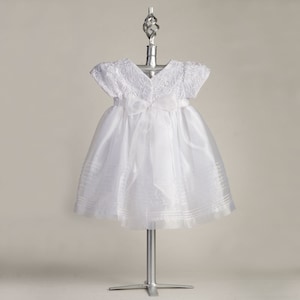 Pu&ograve; includere: Abito bianco per neonata con corpetto in raso e gonna in tulle. L'abito ha un'applicazione di pizzo e un fiocco in vita.