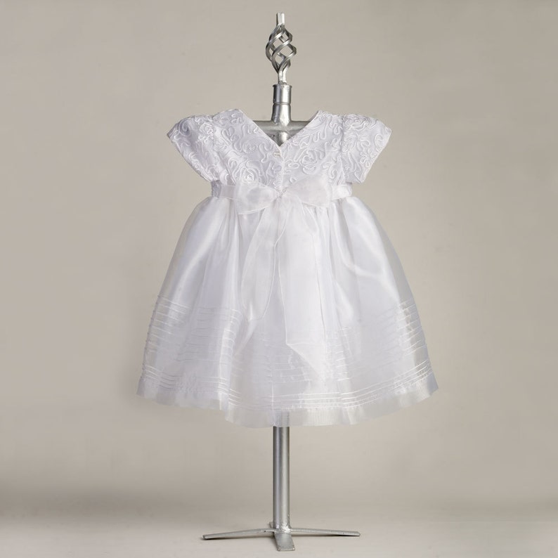 Pu&ograve; includere: Abito bianco per neonata con corpetto in raso e gonna in tulle. L'abito ha un'applicazione di pizzo e un fiocco in vita.