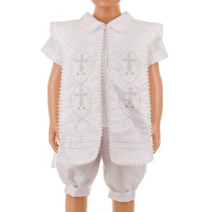 May include: White christening outfit for a baby or young child. The outfit includes a short-sleeved shirt with a collar and a pair of shorts. The shirt has a decorative design with embroidered crosses.