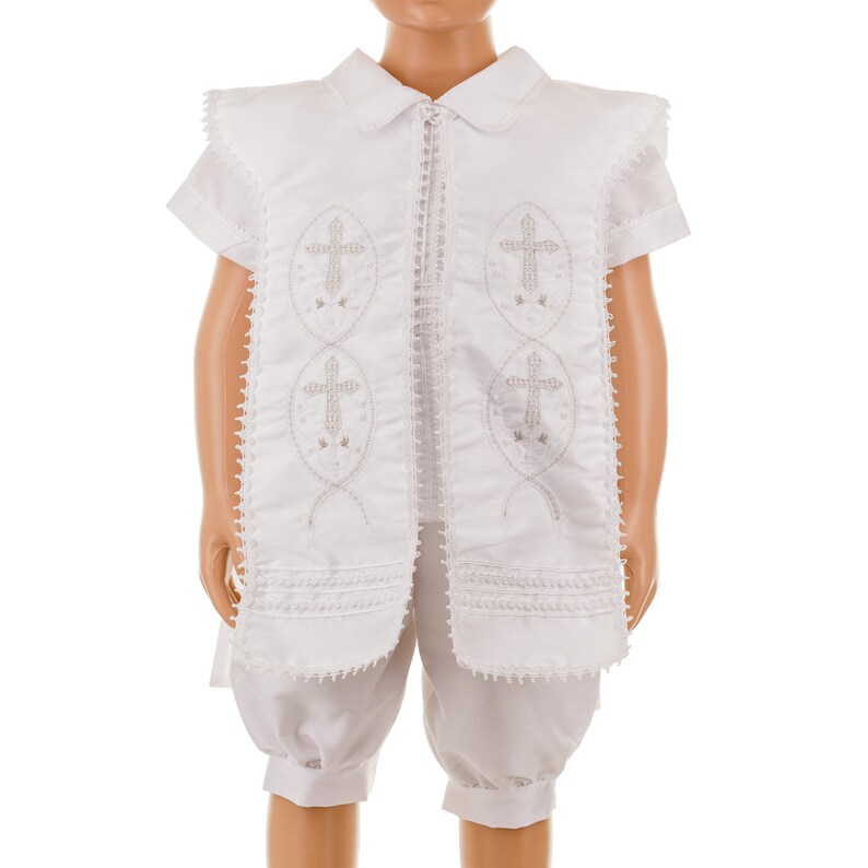 May include: White christening outfit for a baby or young child. The outfit includes a short-sleeved shirt with a collar and a pair of shorts. The shirt has a decorative design with embroidered crosses.