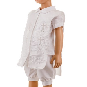 May include: White christening outfit for a baby boy. The outfit includes a short-sleeved shirt with a collar and a matching pair of shorts. The shirt has a decorative embroidered design with crosses.