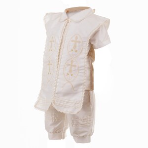 May include: A white christening outfit for a baby or toddler. The outfit includes a short-sleeved shirt with a collar and pants. The shirt has gold embroidered crosses on the front.