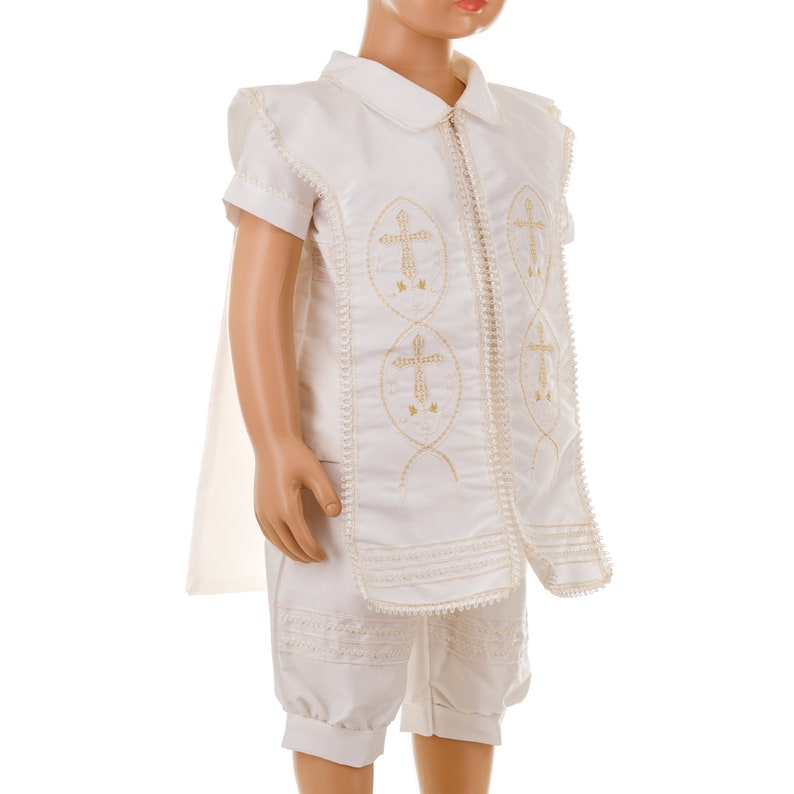 May include: White christening outfit for a baby or young child. The outfit includes a short-sleeved shirt with a collar, a pair of shorts, and a cape. The shirt and shorts have gold embroidered crosses.