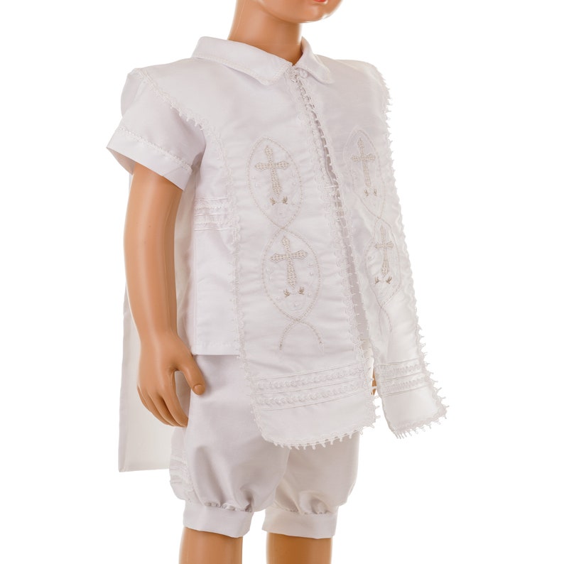 May include: White christening outfit for a baby or young child. The outfit includes a short-sleeved shirt with a collar, a long vest with embroidered crosses, and matching shorts.