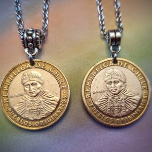May include: Two gold-colored necklaces with silver chains and pendants. The pendants are round coins with the text "REPUBLICA DE CHILE" and "PUEBLOS ORIGINARIOS" around the edges. The center of each coin features a portrait of a person wearing a traditional headdress.