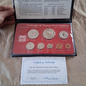 Op de afbeelding: Een 1979 Papua Nieuw-Guinea muntenverzameling in een zwarte presentatiemap met een rode inleg. De set bevat acht munten van verschillende groottes en denominaties, variërend van één toea tot tien kina, inclusief een certificaat van echtheid.