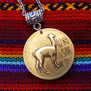 Peut inclure: Un collier avec un pendentif en forme de lama de couleur dorée, avec le texte "UN SOL DE ORO" sur une chaîne en argent avec une perle décorative en argent.